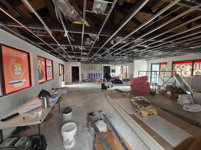 Pinecrest Bakery sales floor mid build-out, with the suspended ceiling grid still open and new ductwork and insulation going in overhead